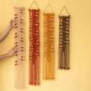 Four macrame wall hangings in beige, rust red, mustard yellow, and dark brown, displayed vertically against a solid pale yellow background. Two hands are visible holding the leftmost beige macrame piece to demonstrate its scale and flexibility.