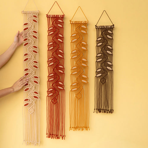 Four macrame wall hangings in beige, rust red, mustard yellow, and dark brown, displayed vertically against a solid pale yellow background. Two hands are visible holding the leftmost beige macrame piece to demonstrate its scale and flexibility.