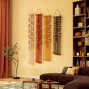 Three bohemian-style woven macrame wall hangings in cream, terracotta, and mustard yellow are displayed on a warm beige wall in a styled living room setting with a brown sofa, wooden bookshelf, and potted plant.