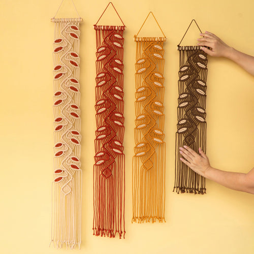 Four bohemian macrame wall hangings with woven seashell motifs are displayed against a solid pale yellow background. Two hands are shown holding the rightmost brown macrame piece to demonstrate its scale and texture.