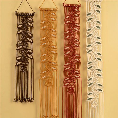 Four vertical macrame wall hangings displayed side by side against a warm beige wall. From left to right, the colors are dark brown, mustard yellow, terracotta red, and off-white, each featuring a braided leaf pattern and hanging from a cord loop at the top.