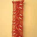 A red macrame wall hanging with a repeating white leaf pattern, suspended against a solid beige background.