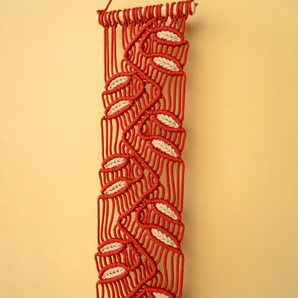 A red macrame wall hanging with a repeating white leaf pattern, suspended against a solid beige background.