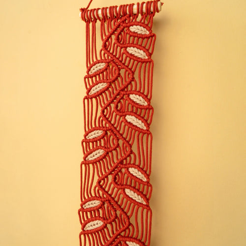 A red macrame wall hanging with a repeating white leaf pattern, suspended against a solid beige background.