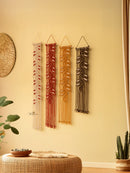 Four macrame wall hangings on a beige wall with a woven ottoman and plants.