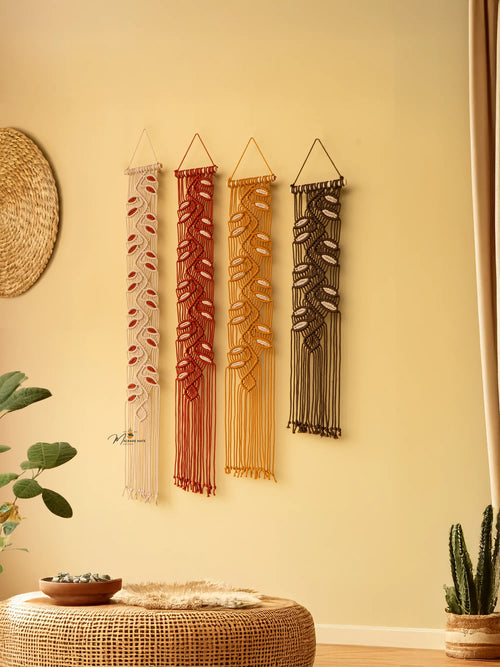 Four macrame wall hangings on a beige wall with a woven ottoman and plants.
