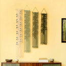 Four vertical macrame wall hangings in cream, sage green, olive green, and black, displayed on a warm yellow wall above a wooden sideboard with decorative bowls and books.