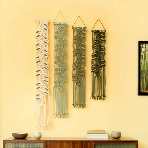 Four vertical macrame wall hangings in cream, sage green, olive green, and black, displayed on a warm yellow wall above a wooden sideboard with decorative bowls and books.