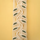 A close-up of a macrame or woven cord trim with a repeating leaf pattern in olive green and cream, set against a solid pale yellow background.