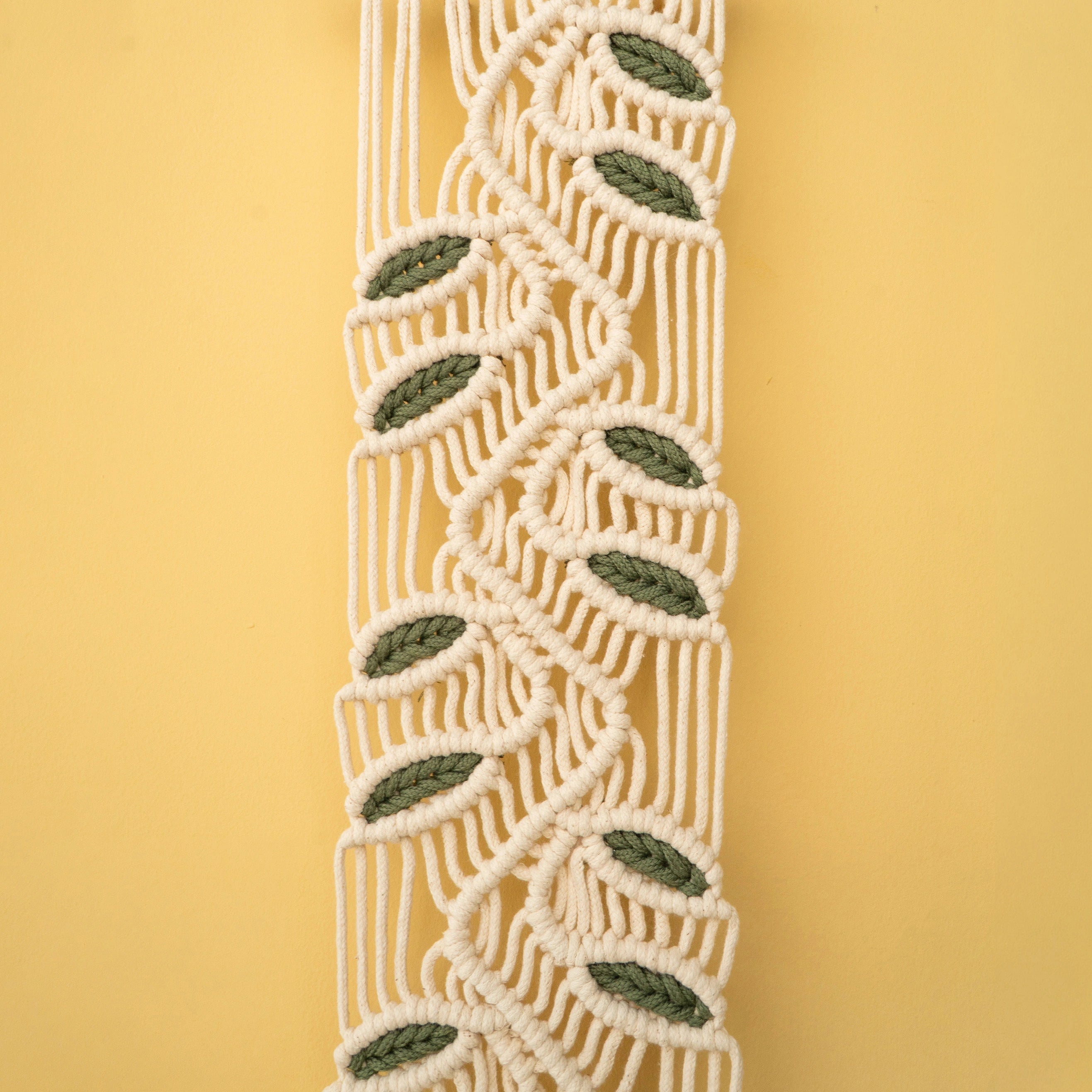 A close-up of a macrame or woven cord trim with a repeating leaf pattern in olive green and cream, set against a solid pale yellow background.