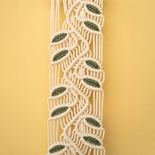 A close-up of a macrame or woven cord trim with a repeating leaf pattern in olive green and cream, set against a solid pale yellow background.