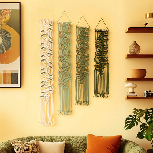 Three bohemian-style macrame wall hangings with leaf patterns in cream, sage green, and dark green, displayed on a warm yellow wall above a green sofa in a styled living room.