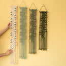 Four macrame wall hangings with leaf patterns in cream, sage green, olive green, and black, displayed on a solid pale yellow wall. Two hands are visible holding the leftmost cream-colored hanging to show scale.