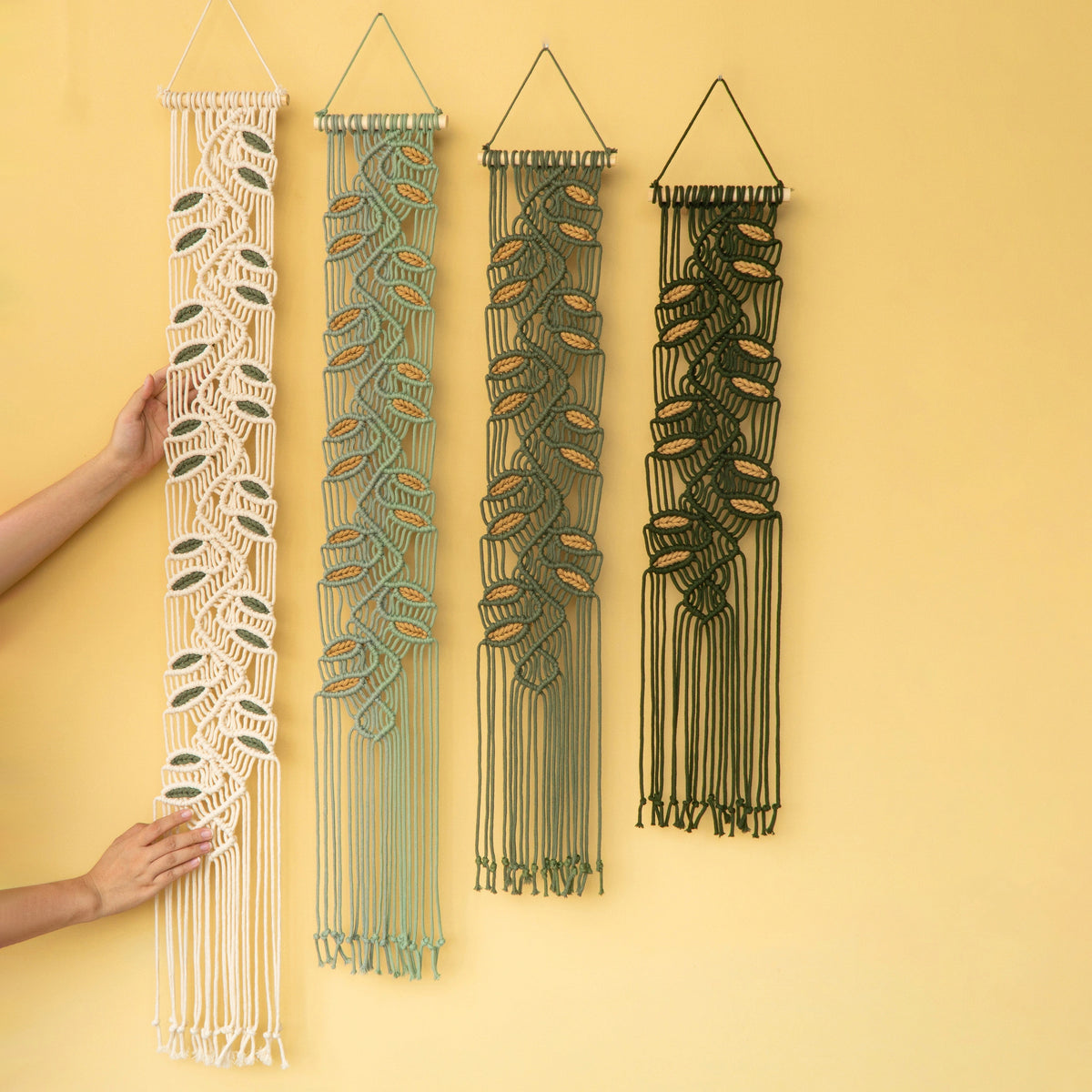Four macrame wall hangings with leaf patterns in cream, sage green, olive green, and black, displayed on a solid pale yellow wall. Two hands are visible holding the leftmost cream-colored hanging to show scale.