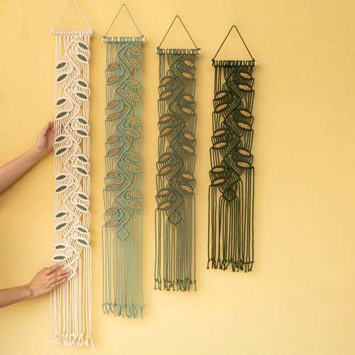 Four macrame wall hangings with leaf patterns in cream, sage green, olive green, and black, displayed on a solid pale yellow wall. Two hands are visible holding the leftmost cream-colored hanging to show scale.