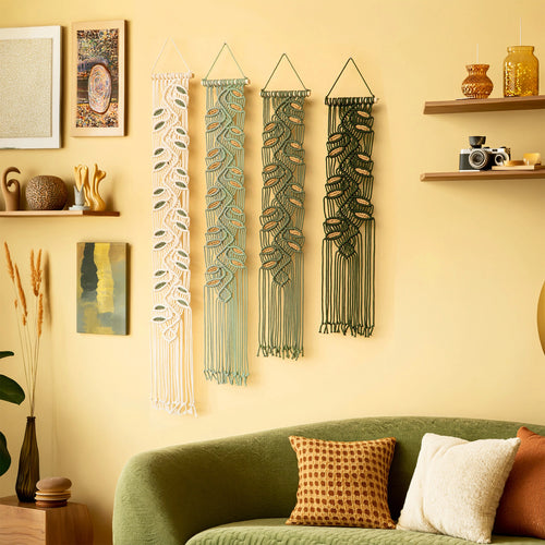 Three woven macrame wall hangings in cream, sage green, and dark green hang on a warm yellow wall above a green sofa with decorative pillows. The room includes shelves with vases and a camera.