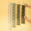 Four woven macrame wall hangings in cream, sage green, dark green, and black, displayed against a solid yellow background. Two hands are shown holding the dark green and black versions to demonstrate scale.
