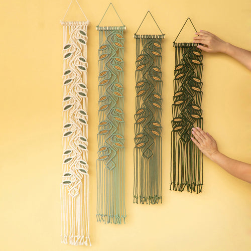 Four woven macrame wall hangings in cream, sage green, dark green, and black, displayed against a solid yellow background. Two hands are shown holding the dark green and black versions to demonstrate scale.