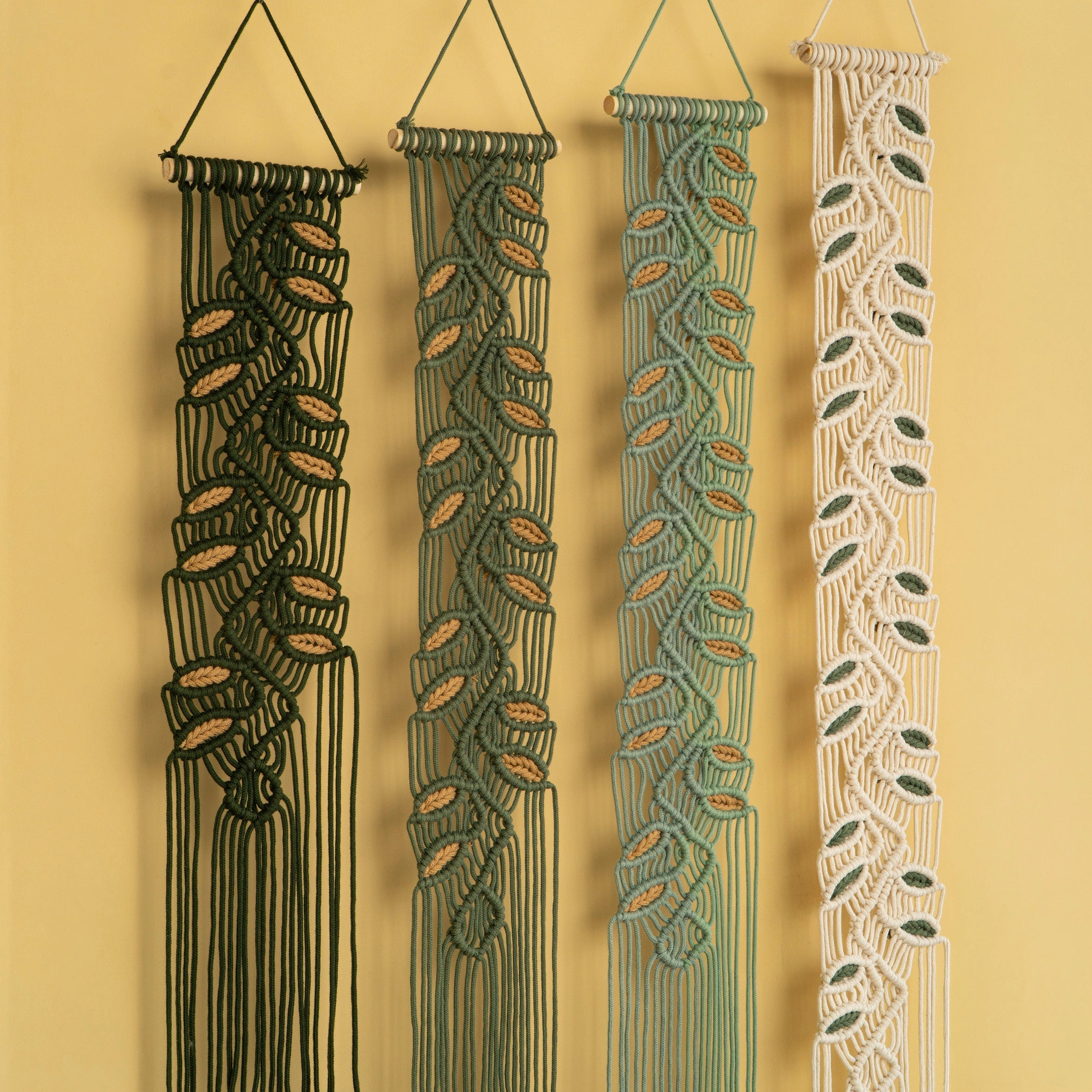 Four macrame wall hangings with leaf-like woven patterns, shown hanging against a warm yellow wall. The hangings are in black, dark green, sage green, and cream, each with a triangular top loop and long fringe at the bottom.