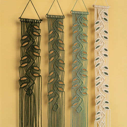 Four macrame wall hangings with leaf-like woven patterns, shown hanging against a warm yellow wall. The hangings are in black, dark green, sage green, and cream, each with a triangular top loop and long fringe at the bottom.