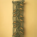 A vertical macrame wall hanging with a leaf pattern, made of dark green cord and tan wooden dowels, shown against a solid beige background.