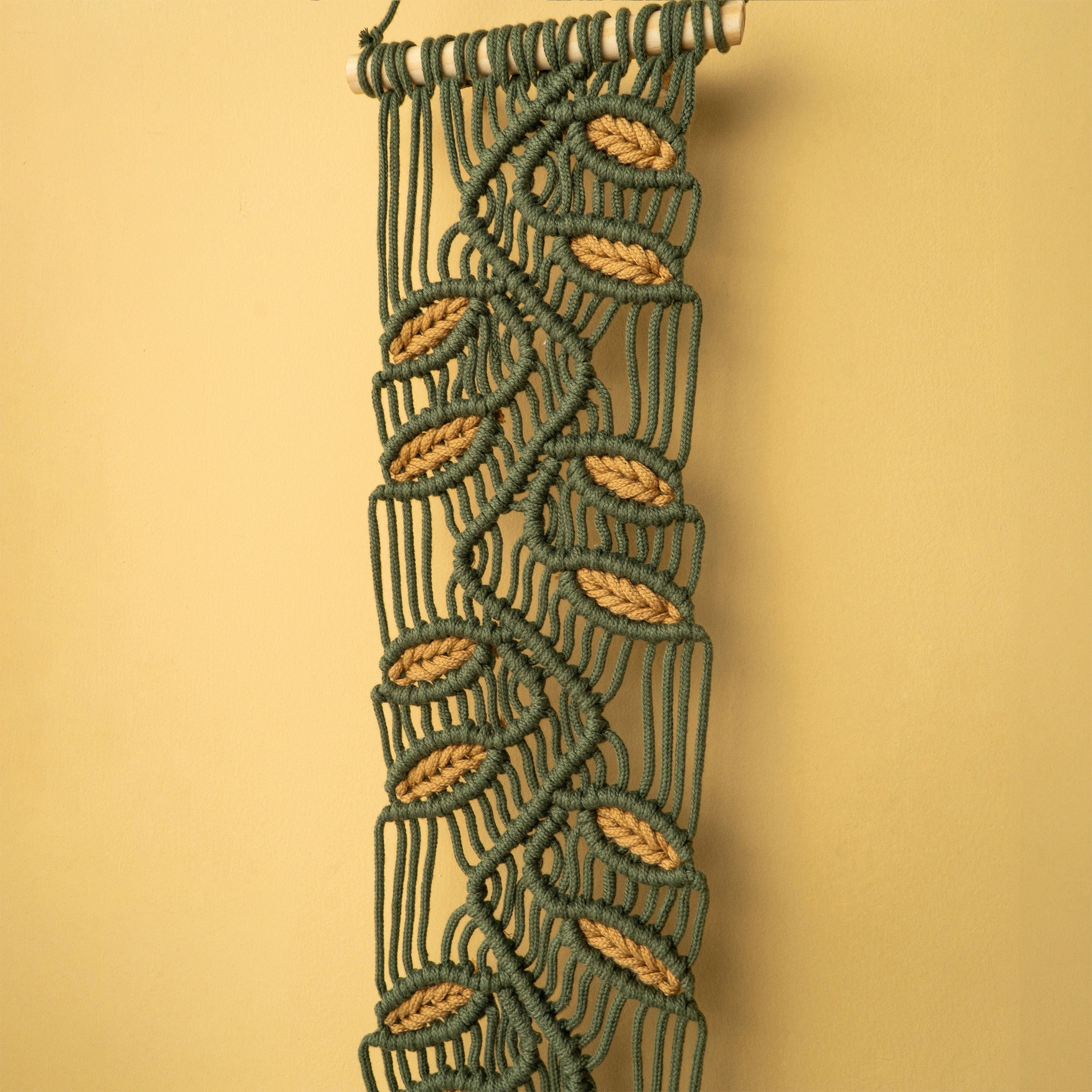 A vertical macrame wall hanging with a leaf pattern, made of dark green cord and tan wooden dowels, shown against a solid beige background.