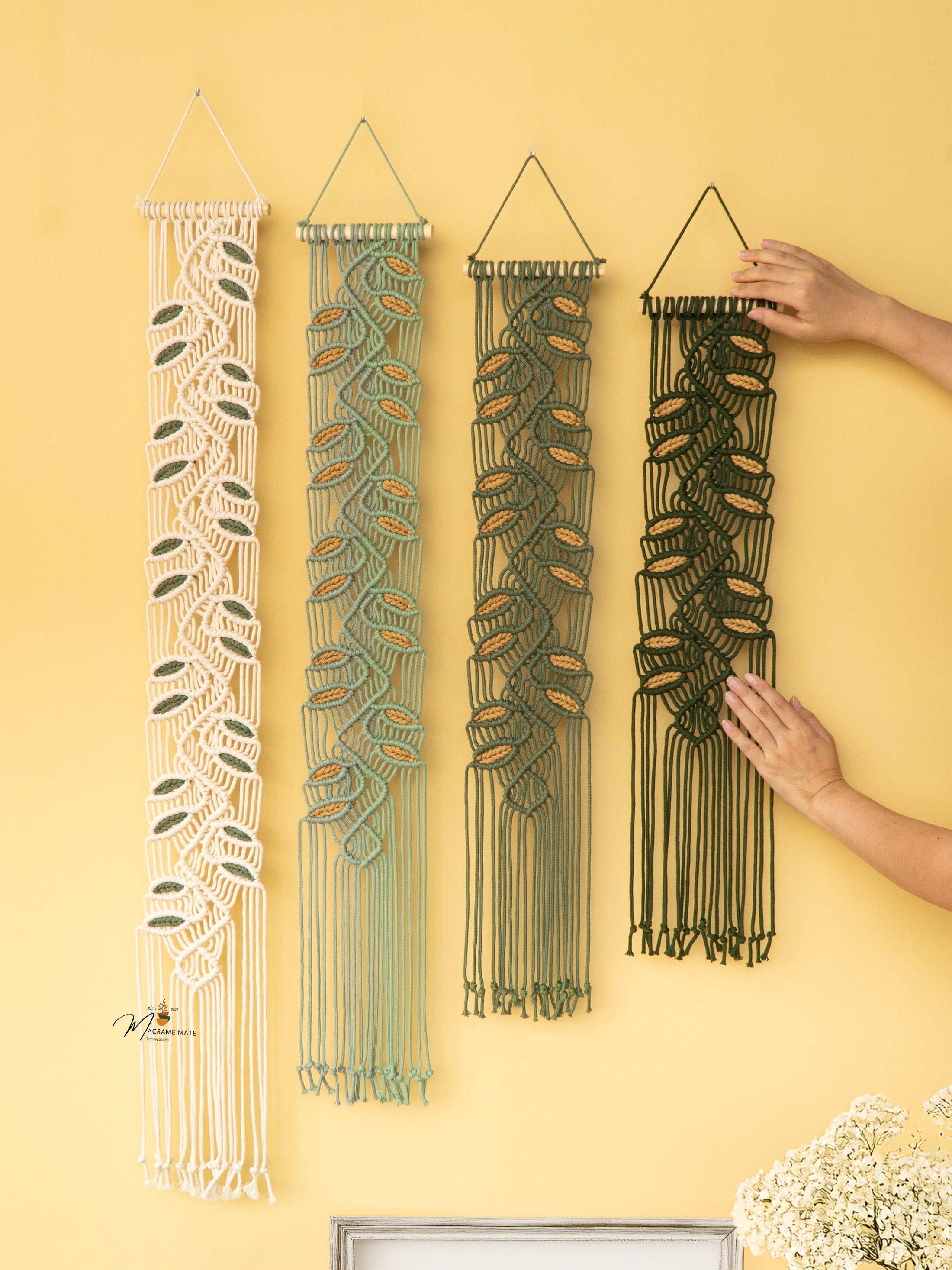 Four woven wall hangings on a yellow wall with a hand reaching for one of them.