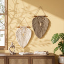 Two bohemian macrame wall hangings in cream and taupe hanging on a warm yellow wall above a wooden cabinet, with a potted plant and books nearby.