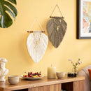Two decorative macrame wall hangings hang on a warm yellow wall above a wooden console table. One hanging is white with a wooden dowel and beads, and the other is taupe-brown with a similar design.