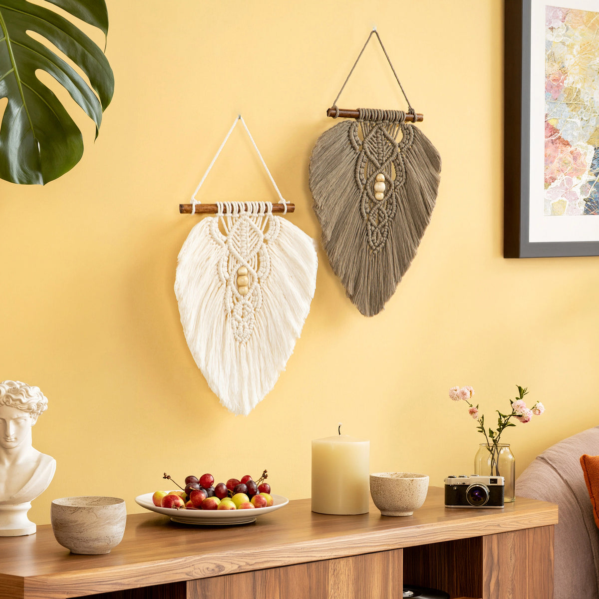Two decorative macrame wall hangings hang on a warm yellow wall above a wooden console table. One hanging is white with a wooden dowel and beads, and the other is taupe-brown with a similar design.