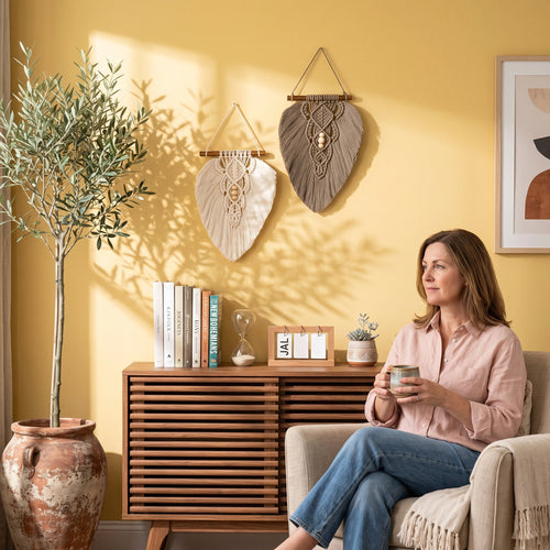 A woman sits in a beige armchair in a warm, sunlit room with yellow walls, holding a mug. Two woven wall hangings are displayed above a wooden cabinet, alongside a large potted olive tree and framed wall art.