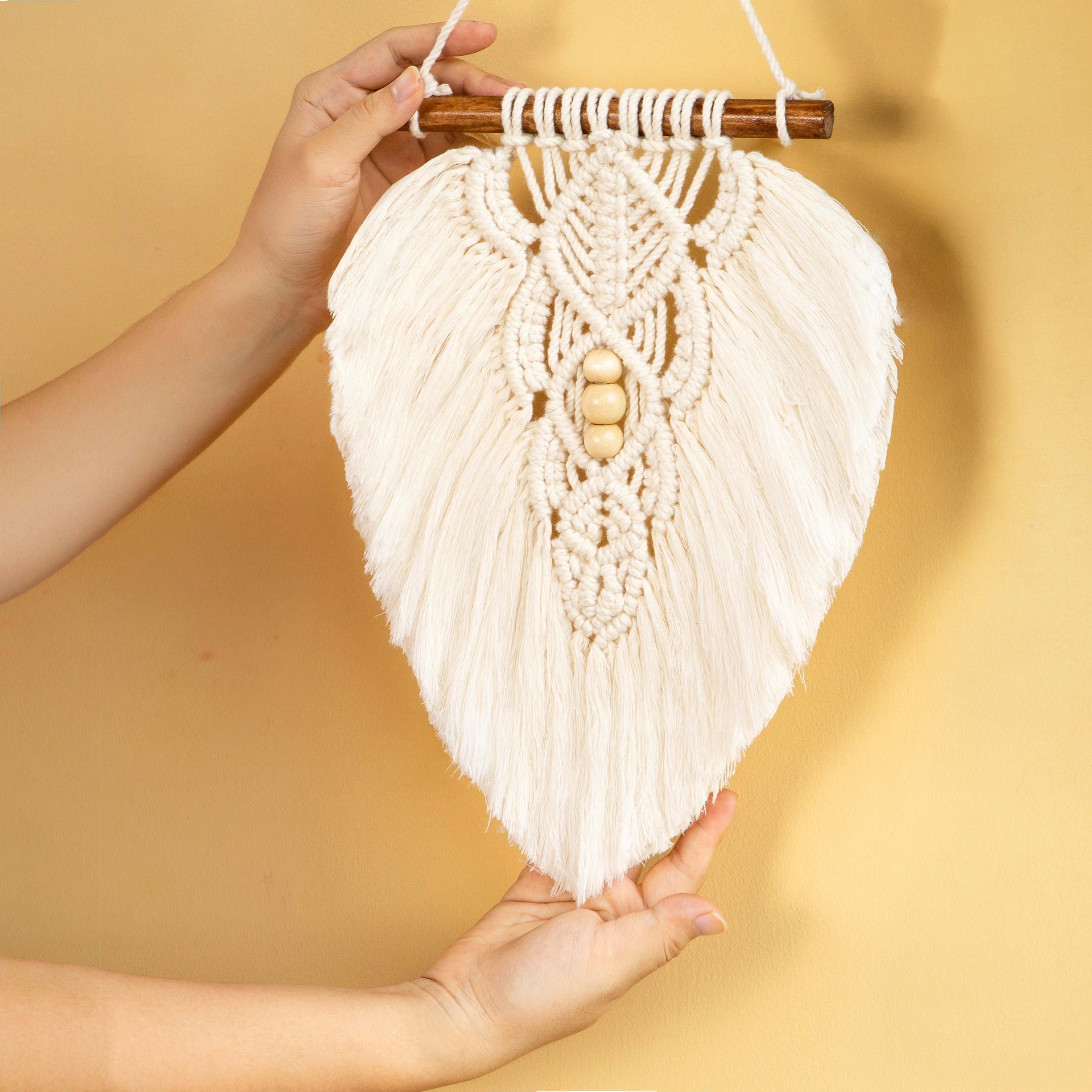 A heart-shaped macrame wall hanging with cream-colored woven cord, a wooden dowel at the top, and three small wooden beads in the center, held up by two hands against a warm beige background.