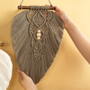 A handmade macrame wall hanging in taupe-brown, suspended from a wooden dowel and held by two hands against a warm beige wall. The central design features three small white wooden beads.
